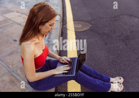 Une femme est assise sur le trottoir en train de taper sur son ordinateur portable ouvert devant elle Banque D'Images