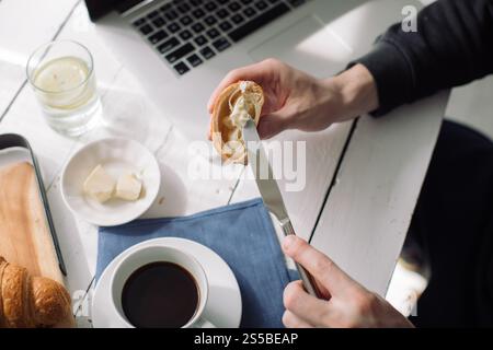 Vue aérienne d'un homme beurrant un croissant et buvant du café tout en utilisant un ordinateur portable Banque D'Images