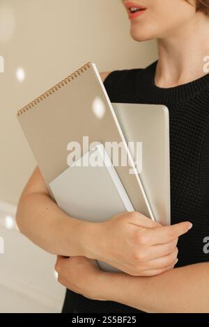 Gros plan d'une jeune femme dans une robe noire élégante debout près d'un mur transportant un ordinateur portable, un bloc-notes et un carnet de notes Banque D'Images
