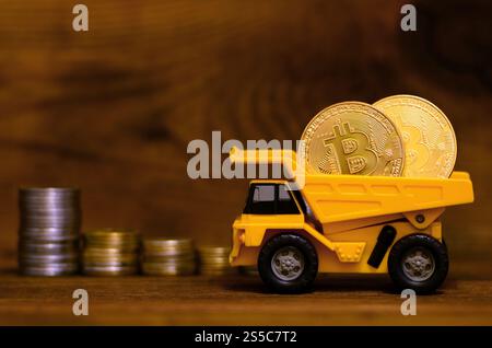 Le dumper de jouet jaune chargé de bitcoins dorés brillants se tient à côté de piles de pièces de monnaie de différentes devises sur un fond en bois. L'avantage Banque D'Images