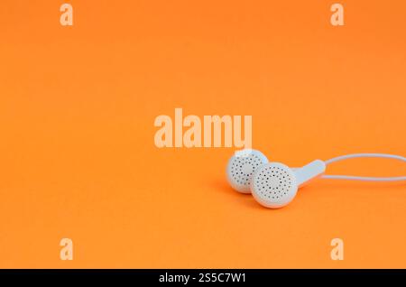 Les écouteurs modernes blancs reposent sur un fond orange vif. Un modèle pour les fans d'écoute de musique. Écouteurs modernes blancs repose sur un orange vif Banque D'Images