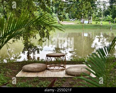 Un salon extérieur confortable avec une table tissée et des coussins sur une natte à côté d'un lac paisible Banque D'Images