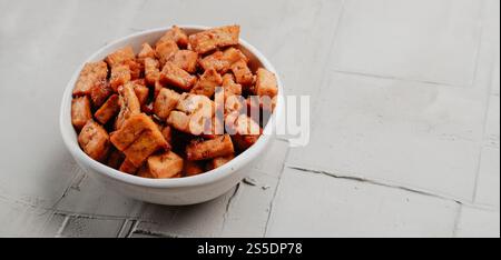 quelques cubes de tofu marinés, servis comme collation sur un bol en céramique blanche, sur une surface en béton rustique, dans un format panoramique à utiliser comme bannière web Banque D'Images