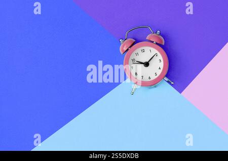 Un petit réveil rose est situé entre les zones de couleurs bleu, violet et rose. Concept de gestion du temps. Modèle minimal de pose à plat avec copie Banque D'Images