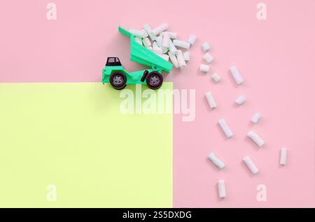 Green Little Toy dump truck jette des morceaux de guimauve de son dos soulevé sur un fond jaune pastel et corail. Vue de dessus minimale à plat. Vidage Banque D'Images