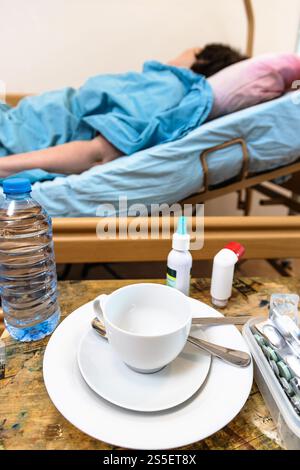 vider la tasse avec soucoupe, les médicaments et une bouteille d'eau sur la table et le patient dans le lit d'hôpital à domicile sur le fond à la maison Banque D'Images