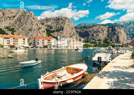 Paysage marin ensoleillé avec des bateaux amarrés le long d'une baie calme dans la rivière Cetina flanquée d'imposantes falaises et de bâtiments sous un ciel bleu avec des nuages moelleux, omis, Croatie Banque D'Images