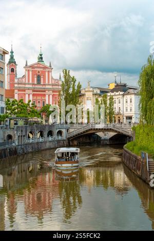 Un bateau d'excursion glisse sous un pont de pierre orné sur la rivière Ljubljana, avec des bâtiments historiques aux couleurs pastel et l'église franciscaine de l'Annonciation en toile de fond sous un ciel nuageux, Ljubljana, Slovénie Banque D'Images