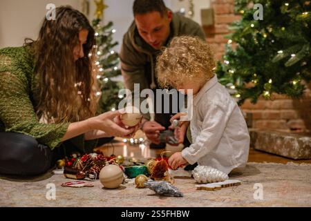 Famille décorant ensemble devant un sapin de Noël scintillant, avec un enfant concentré sur les boules et les ornements, Ohio, États-Unis Banque D'Images