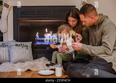 Une famille profite d'un moment agréable au coin de la cheminée, manger des biscuits et boire du lait pendant Noël, Ohio, États-Unis Banque D'Images