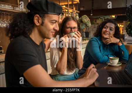 Trois amis profitent d'une conversation autour d'un café-bar confortable, travaillant sur un ordinateur portable, Berlin, Allemagne Banque D'Images