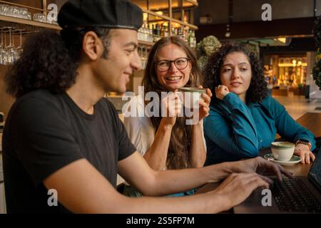 Trois amis profitent d'une conversation autour d'un café-bar confortable, travaillant sur un ordinateur portable, Berlin, Allemagne Banque D'Images
