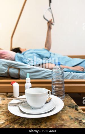 tasse avec soucoupe et bouteille d'eau sur la table et malade tenant la poignée du lit d'hôpital de soins à domicile sur le fond à la maison Banque D'Images