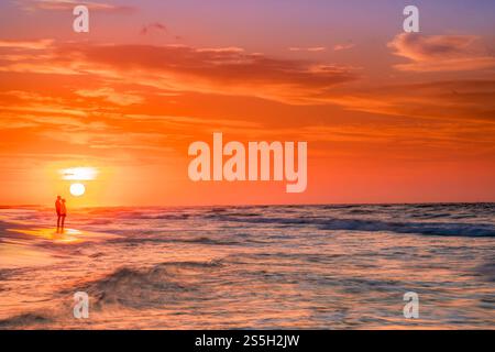 Un couple se tient ensemble le long de la côte à la plage au coucher du soleil, silhouetté contre la lumière chaude du soir et l'horizon océanique incandescent. Banque D'Images