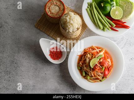 Salade de papaye thaïlandaise dans une assiette blanche avec riz collant dans un panier en osier bambou et crevettes séchées. Banque D'Images