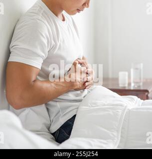 Un homme est assis au lit avec un mal d'estomac et presse son estomac avec ses mains. Banque D'Images