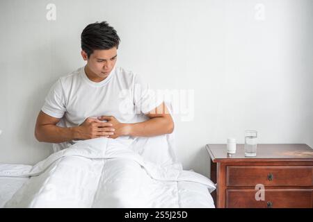 Un homme est assis au lit avec un mal d'estomac et presse son estomac avec ses mains. Banque D'Images