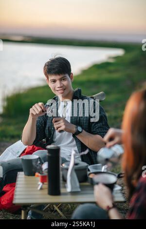 Mise au point sélective, bel homme faisant moulin à café frais, joyeux couple jeune routard assis devant la tente près du lac avec set de café Banque D'Images