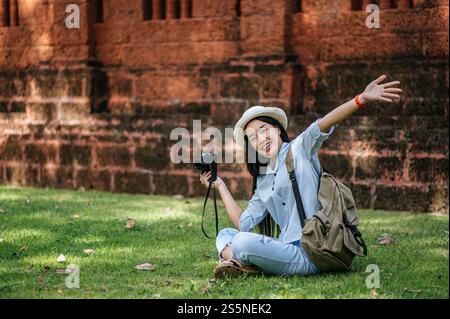 Jeune routard joyeux assis sur l'herbe levez son bras et souriant avec bonheur, tenant un appareil photo à la main tout en voyageant dans un site antique Banque D'Images