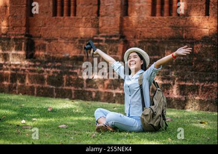 Jeune routard joyeux assis sur l'herbe levez son bras et souriant avec bonheur, tenant un appareil photo à la main tout en voyageant dans un site antique Banque D'Images