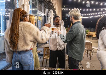 Groupe de jeunes amis multiethniques grillant debout sur une terrasse de bar la nuit Banque D'Images