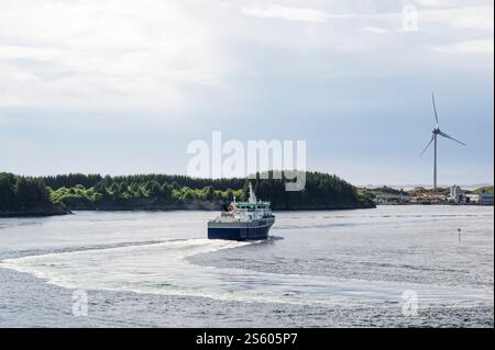 Un bateau de travail, naviguant au large des côtes du sud de la Norvège, avec une éolienne visible sur le rivage Banque D'Images