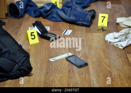Enquête sur les lieux du crime - numérotation des preuves après le meurtre dans l'appartement. Smartphone cassé, portefeuille et vêtements avec marqueurs de preuves Banque D'Images