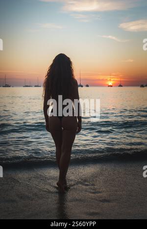 Une femme aux cheveux bouclés fait face à un coucher de soleil vibrant sur les vagues calmes de l'océan, avec des voiliers silhouettes à l'horizon dans les îles Phuket ou Koh Phi Phi. Banque D'Images