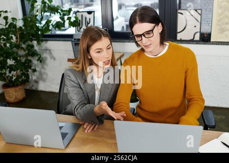 Deux jeunes personnes s'engagent dans une discussion animée tout en travaillant sur des ordinateurs portables dans un espace de travail élégant. Banque D'Images