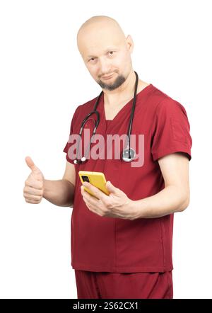 Un médecin chauve portant des gommages rouges et un stéthoscope autour du cou sourit tout en tenant un smartphone jaune. Il fait un geste du pouce levé Banque D'Images