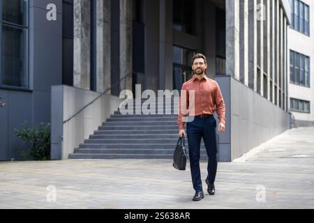 Un homme d'affaires prospère en pleine croissance se promène dans la ville avec un sac d'affaires dans ses mains, un homme mature sourit avec satisfaction de l'extérieur d'un immeuble de bureaux. Banque D'Images