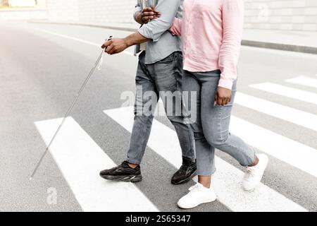 Vue rognée de la femme soignante aidant un homme noir malvoyants avec canne à travers la rue de ville, de plus près Banque D'Images