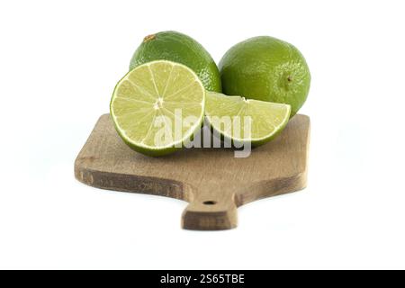 Avec deux citrons verts entiers à côté d'un citron vert tranché et d'un coin de citron vert, le tout disposé sur une petite planche à découper en bois sur un fond blanc propre Banque D'Images