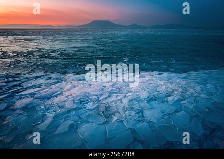 Fonyod, Hongrie - vue panoramique sur le lac Balaton gelé par un après-midi froid d'hiver avec glace brisée, ciel de coucher de soleil coloré doré et le mont Badacsony Banque D'Images