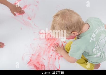 Bébé joyeusement peinture au doigt avec une couleur rouge vif sur une grande feuille de papier blanc, s'engageant dans un jeu désordonné tout en favorisant la créativité et de sensoriel de Banque D'Images