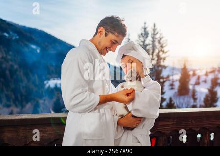 Jeune couple tenant chat sur le balcon de l'hôtel profitant du paysage de montagne d'hiver portant des peignoirs. Voyagez avec votre animal de compagnie Banque D'Images