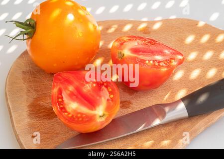 Fermez les tomates rouges et jaunes et un couteau de cuisine sur une planche de bois sur un fond blanc. Ombres abstraites, vue de dessus. concept d'été et de végéta Banque D'Images