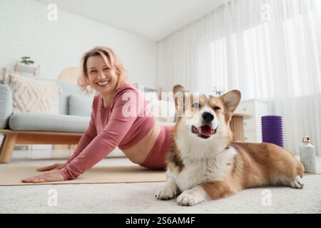 Belle femme sportive mature heureuse pratiquant le yoga avec un chien mignon Corgi à la maison Banque D'Images