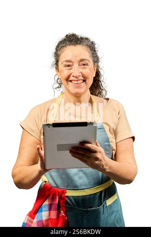 Portrait d'une femme mature et joyeuse portant un tablier et utilisant une tablette numérique, éventuellement en suivant une recette ou en gérant des commandes Banque D'Images