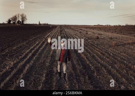 Image aérienne d'un agriculteur senior avec un ordinateur portable dans une main faisant signe avec l'autre pour saluer quelqu'un dans le champ après le processus de mise en balles à la fin de l'automne Banque D'Images