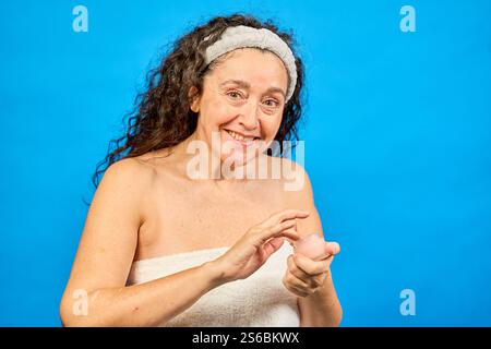 Femme d'âge moyen portant une serviette de bain appliquant la crème pour le visage après la douche, souriant et regardant la caméra sur fond bleu Banque D'Images