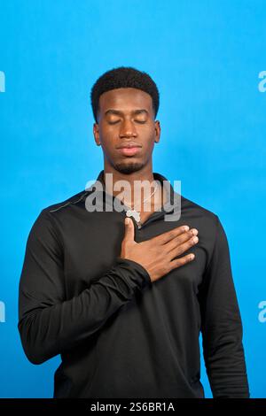 Jeune sportif avec les yeux fermés, tenant la main sur le cœur sur fond bleu, montrant du respect, du patriotisme ou commémorant un événement important Banque D'Images