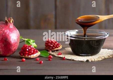 Mélasse de grenade dans un bol en verre, sauce aigre à la grenade avec des fruits de grenade entiers et fendus frais mûrs sur une table rustique en bois (Punica granatum) Banque D'Images