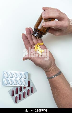 Femme âgée versant des pilules d'une bouteille brune dans sa main, avec des blisters de capsules rouges et blanches à proximité, mettant l'accent sur les soins à domicile Banque D'Images
