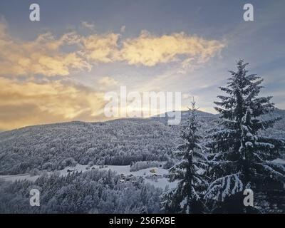 Paysage hivernal serein avec des conifères couverts de neige sur les collines sous un ciel de coucher de soleil. Les maisons nichées dans les bois créent une scène pittoresque. Banque D'Images