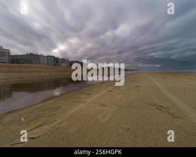 Un paysage de plage pittoresque avec un plan d'eau réfléchissant, des bâtiments modernes adjacents et des nuages couverts spectaculaires. Capture la tranquillité et la sére Banque D'Images