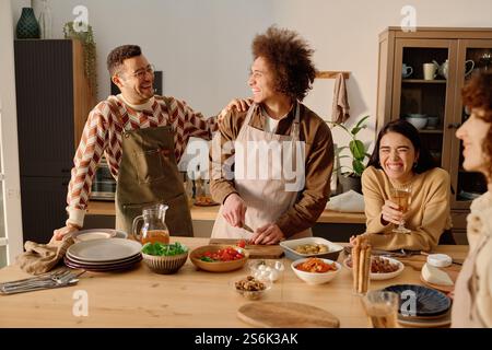 Souriant jeune homme caucasien adulte avec les cheveux courts plaisantant et tapotant ami biracial aux cheveux bouclés sur l'épaule tout en coupant la dernière fois des tomates, jeune femme adulte appuyée sur la table et riant à voix haute Banque D'Images