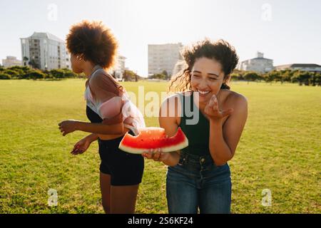 Un moment ludique entre deux jeunes amis riant et mangeant de la pastèque lors d'une journée ensoleillée au parc dans leur ville, profitant de l'amitié et de la HAP Banque D'Images