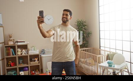 Jeune homme prenant selfie dans une chambre avec lit bébé, souriant et entouré de jouets pour bébés, créant une ambiance chaleureuse et familiale dans un cadre intérieur moderne. Banque D'Images