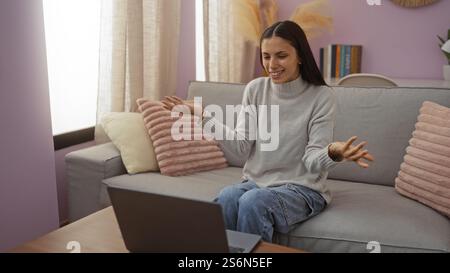 Femme ayant un appel vidéo dans un cadre de salon confortable, s'engageant joyeusement tout en étant assise sur un canapé avec des coussins roses et des décorations d'intérieur autour d'elle Banque D'Images